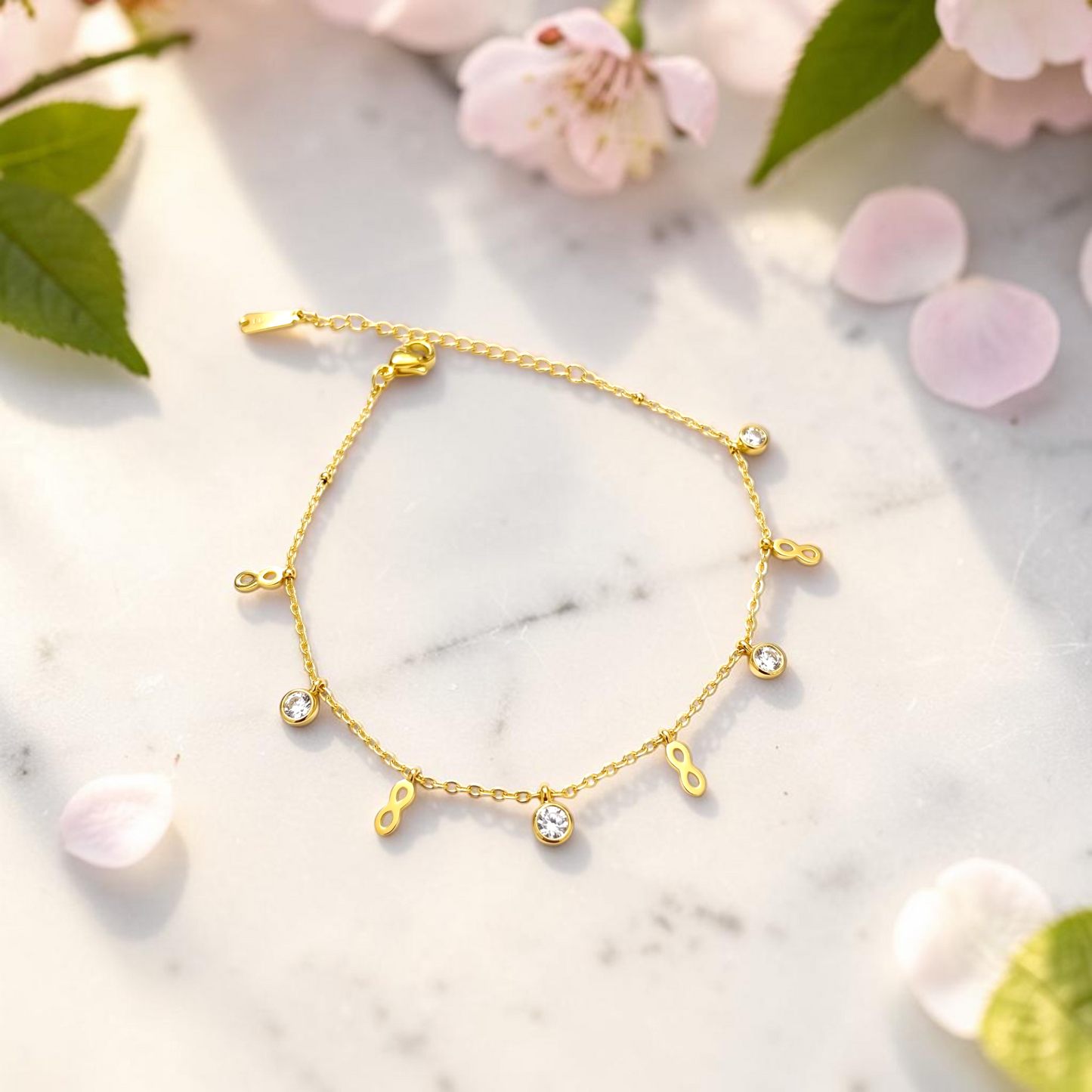 AVIRI gold-tone infinity charm bracelet with clear cubic zirconia stones on a marble surface with flower petals.