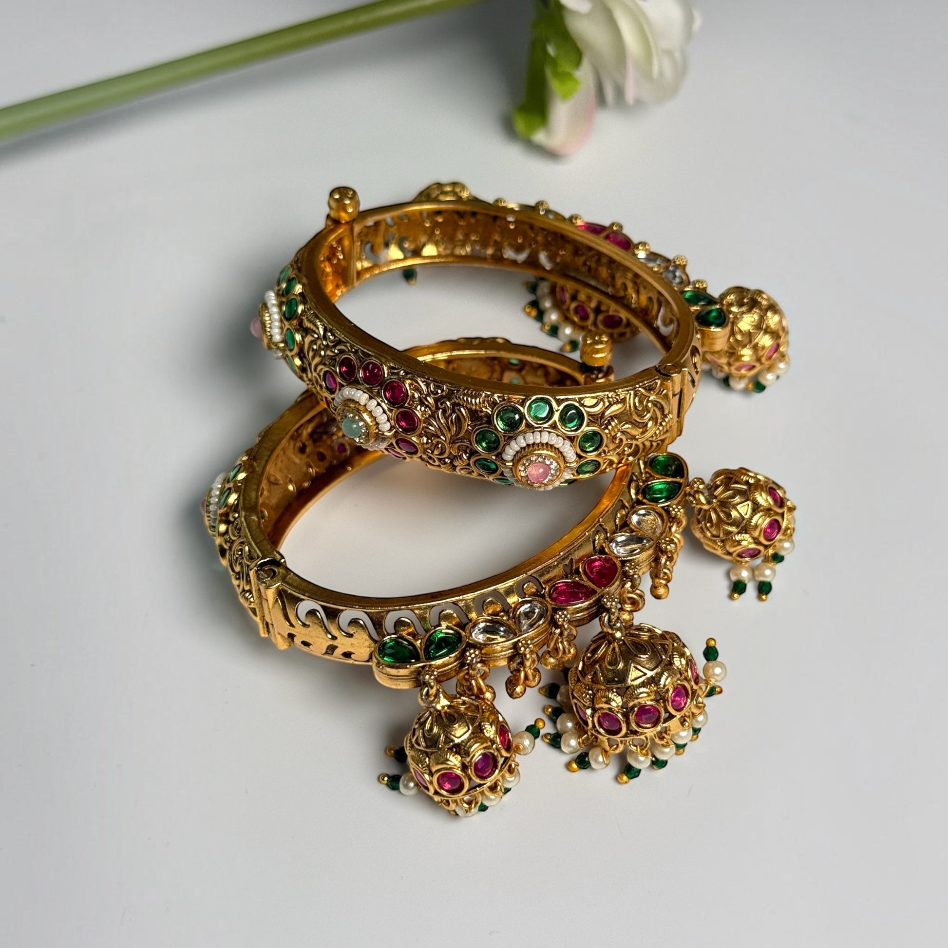 AVIRI traditional gold-tone hinged bangle bracelets with red and green Kundan stones and dangling jhumka charms.