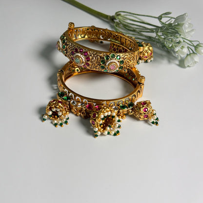 AVIRI gold-tone hinged bangle bracelets with red and green Kundan stones, floral filigree, and dangling jhumka charms.