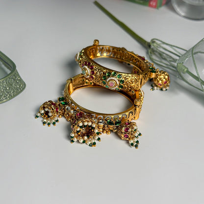 AVIRI traditional gold-tone hinged bangles with red and green Kundan stones, floral filigree, and dangling jhumka charms.