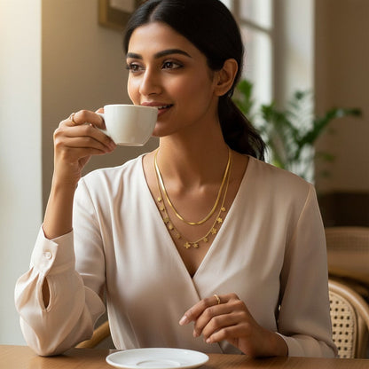 AVIRI gold-tone layered necklace with herringbone chain and clover charms. Model wears it with a cream blouse.